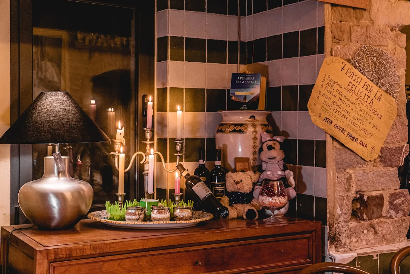 Trattoria sideboard with a lamp, lit candles, bottles and details of stone and tiled walls.