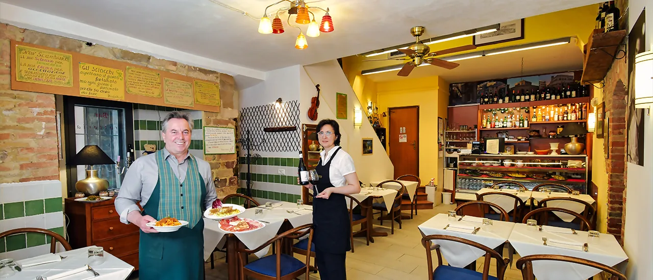 Lolita and Vittorio in the Bottega Dei Priori dining room, holding dishes prepared in the kitchen and a bottle of wine.